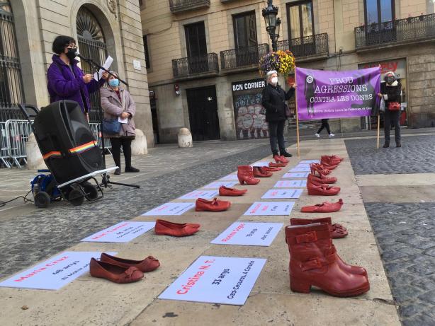 Fotografi on apareix en primera plana 19 parells de sabates vermelles amb els noms de les dones assassinades el 2021 i al fons, una dona parlant al micròfon.